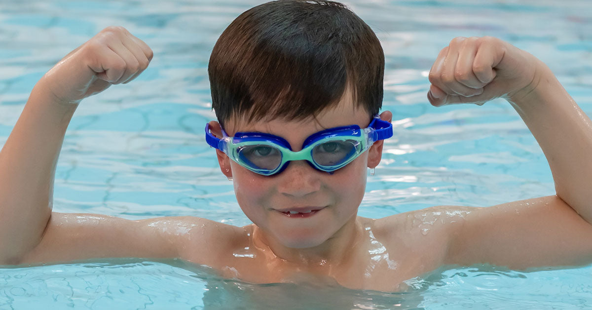 Confident young boy flexing arms while wearing blue kids swimming goggles in a swimming pool.