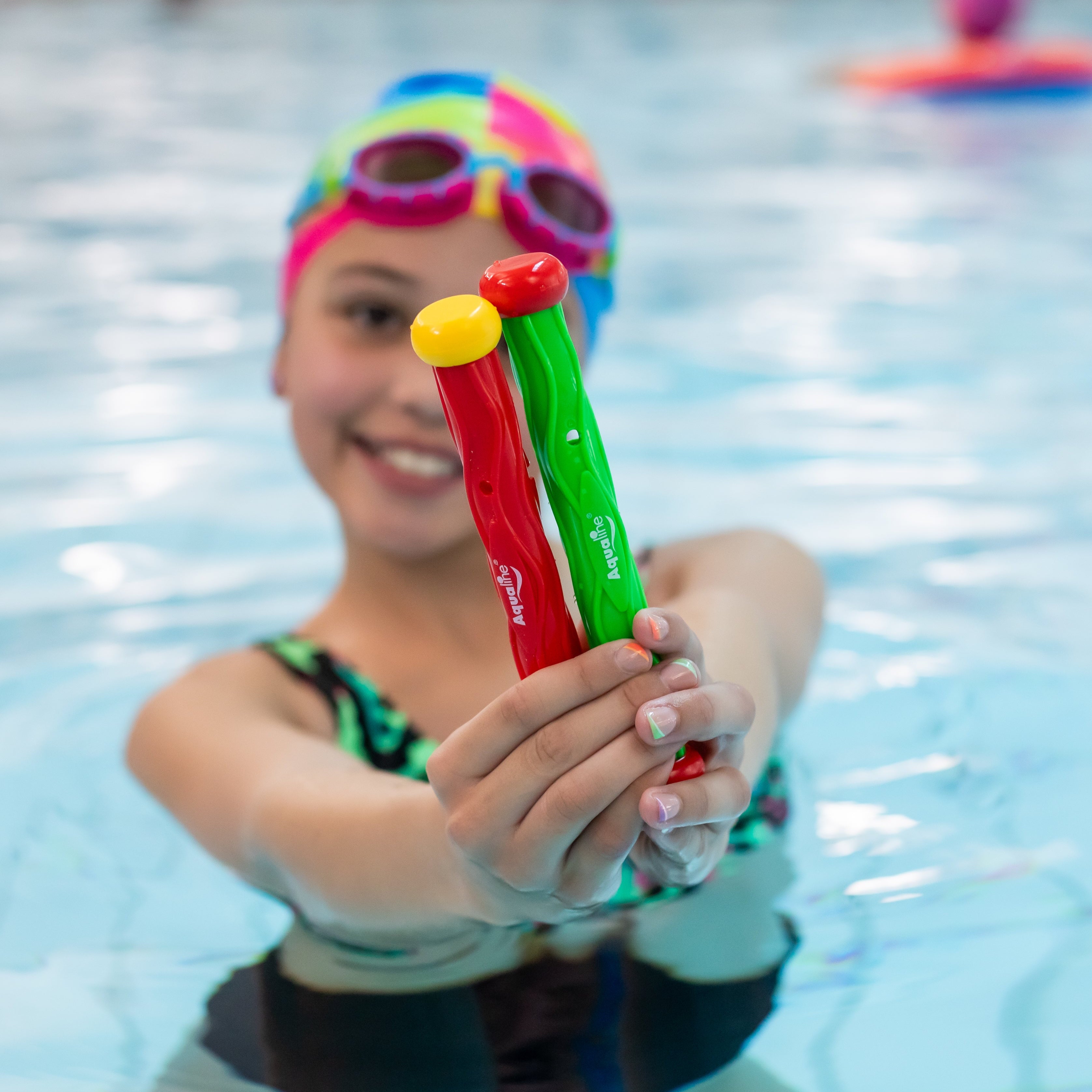 Child in a pool holding colorful swim dive sticks with a swim cap and goggles.