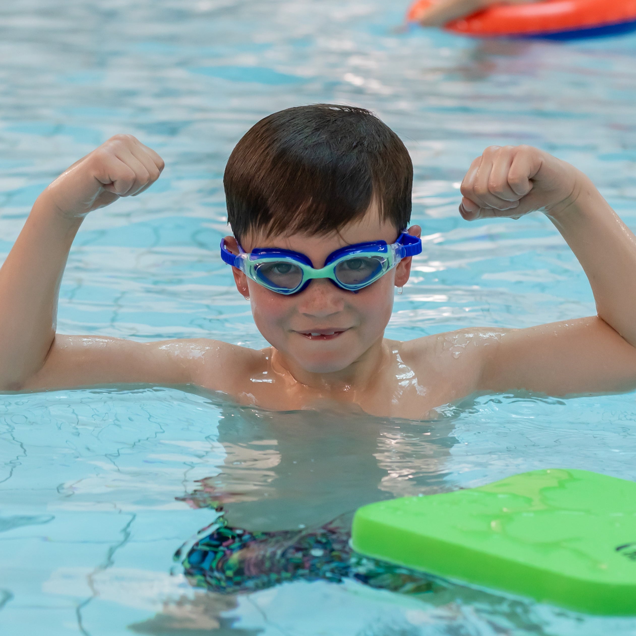 Child wearing goggles flexing muscles in a pool with a green floatation device nearby