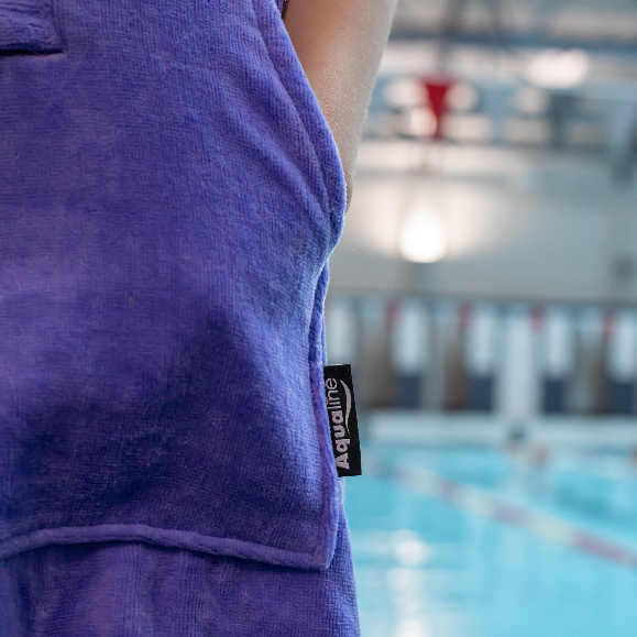Person wearing a purple towel with Aqualine branding by an indoor pool.