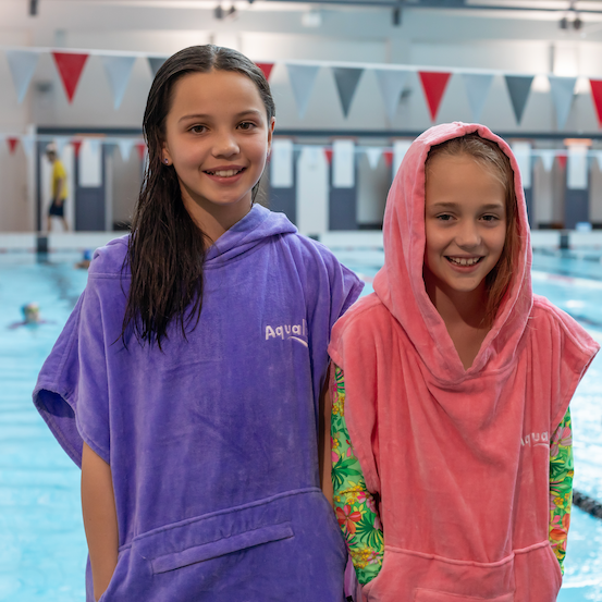 Two children wearing purple and pink hooded towels by a pool.